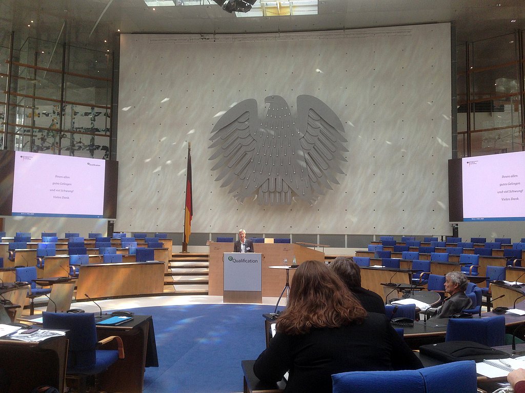 EIn Saal, im Hintergrund der Bundesadler, vorne ein Sprecher auf dem Podium